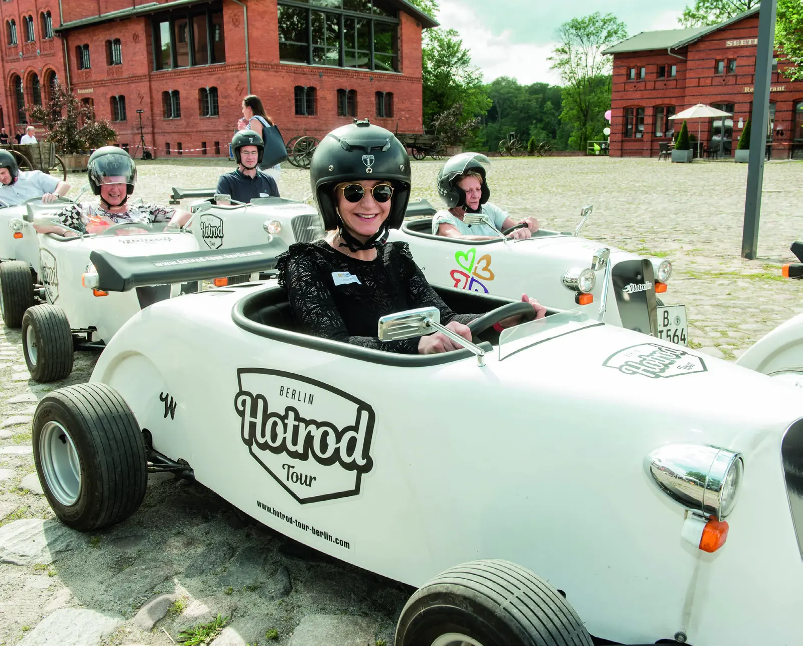 Teilnehmende einer Hotrod-Tour auf einem Hof aus Backsteingebäuden