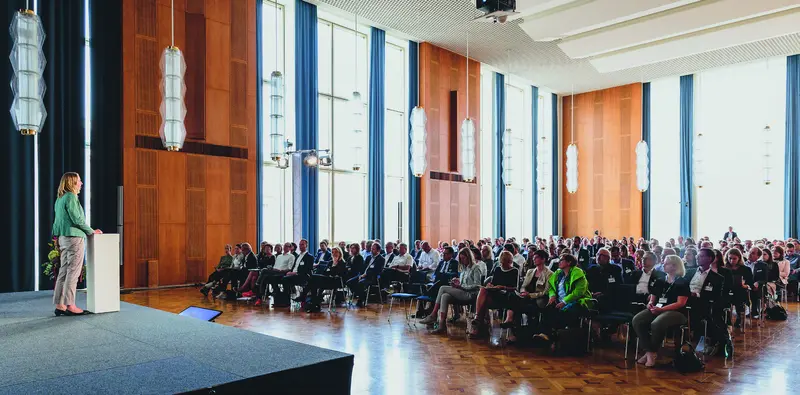 Tagung in einem holzvertäfelten Saal mit Rednerin am Pult