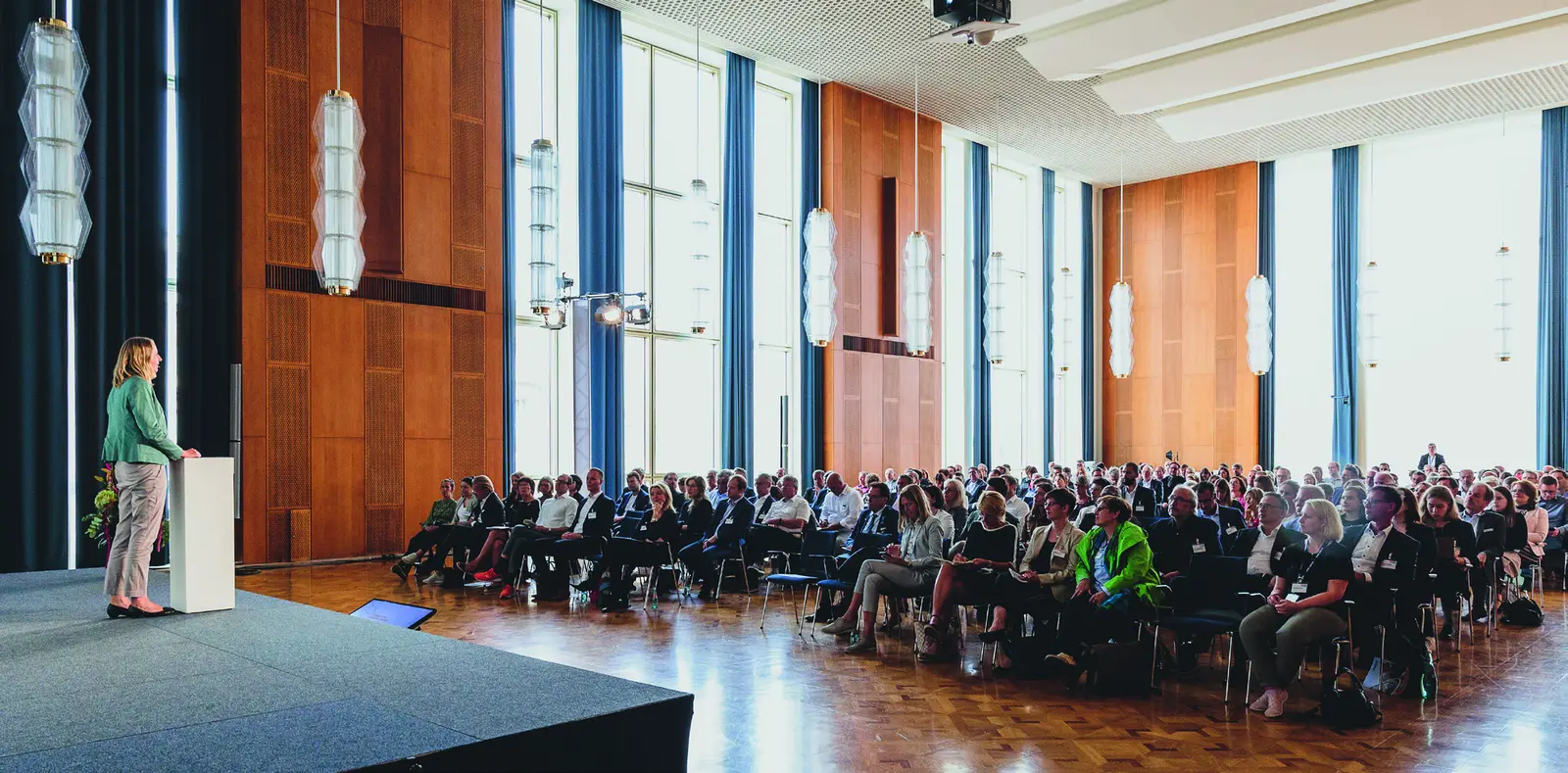 Tagung in einem holzvertäfelten Saal mit Rednerin am Pult